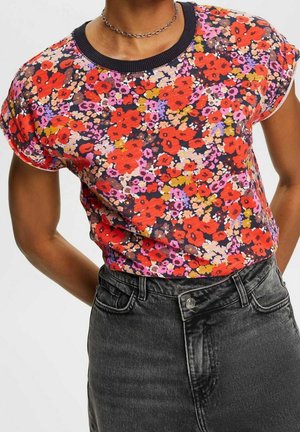 Person iført en kortærmet blomstret top med røde, lyserøde og orange blomster og højtaljede, falmede sorte jeans med en kædehalskæde.