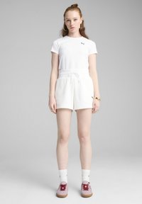 T-shirt bianco a maniche corte con logo nero, abbinato a pantaloni corti bianchi con lacci. Il modello indossa scarpe da ginnastica rosa e calzini bianchi con dettagli neri.