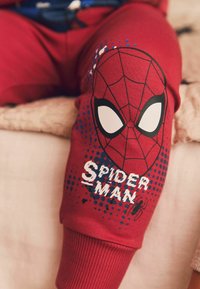 Rote Sweatpants mit einem großen Spider-Man-Masken-Graphic in Schwarzumrandung und weißen Augen, ergänzt durch blaue Punktmuster und Text.