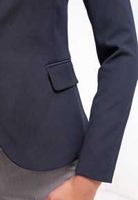 Granatowa marynarka z teksturowanej tkaniny, o dopasowanym kroju i pojedynczej kieszeni z przodu. Warstwowo noszona pod szarym ubraniem.