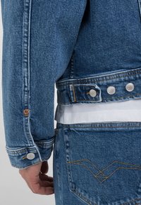 Jeans jakna z modrim pranjem, ki ima srebrne snemalne gumbe, oranžno šivanko in teksturirano tkanino. Zadnji žep ima šivan vzorec.