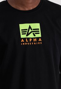 Must sulgeda must t-särk, millel on roheline ristkülikukujuline logo tähisega "A" ja kolm triipu, ning oranž tekst "ALPHA INDUSTRIES."