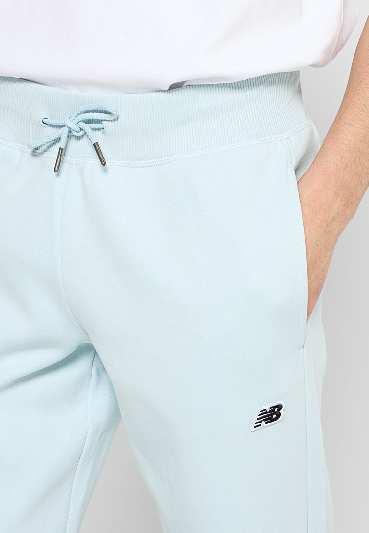 Un pantalon de survêtement bleu clair avec une taille élastique équipée d'un cordon de serrage, une texture côtelée, des poches latérales et un petit logo noir sur la cuisse.