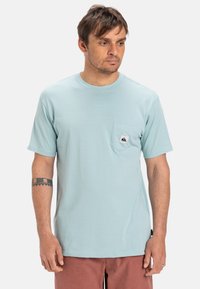 Světle modré tričko s krátkým rukávem vyrobené z měkké látky, s náprsní kapsou s logovou nášivkou. Pravidelný střih a kulatý výstřih.