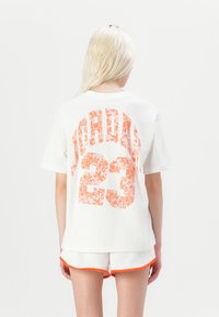 White cotton t-shirt featuring orange floral-patterned graphics reading "JORDAN" and "23" on the back, with short sleeves and a relaxed fit.