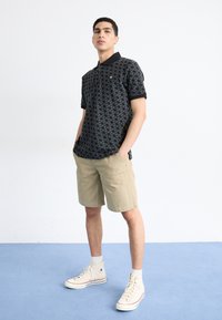 Sort mønstret polo t-shirt med korte ærmer, khaki shorts og beige højhælede sneakers, sat mod en ensfarvet hvid baggrund.