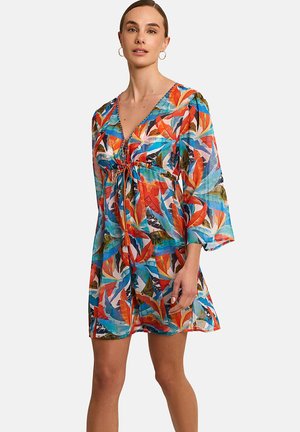 Femme portant une robe ample et colorée avec des motifs de feuilles orange, bleu et vert, et un profond décolleté en V noué à l'avant.
