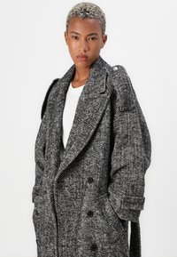Cappotto grigio a spina di pesce con bottoni doppi, ampi risvolti e spalle strutturate. Presenta tasche frontali e una cintura in vita.