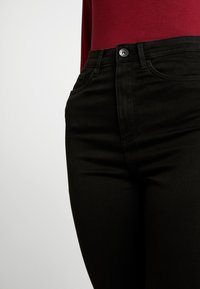 Zwarte spijkerbroek met een slim fit, voorzien van voorzakken, een knoopsluiting en stikseldetails, gecombineerd met een bordeauxrode top met lange mouwen.