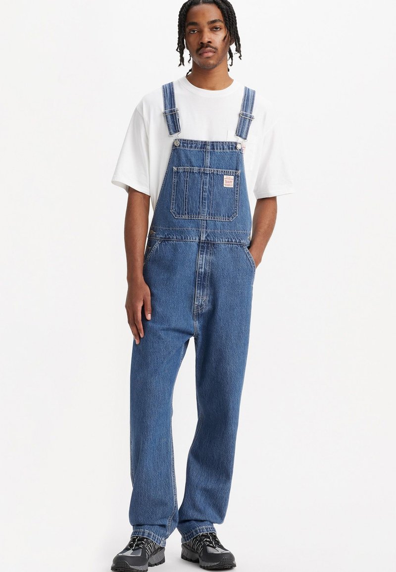 Salopette en denim bleu moyen, avec des jambes larges, des bretelles ajustables et une poche avant avec étiquette. Associée à un T-shirt blanc.