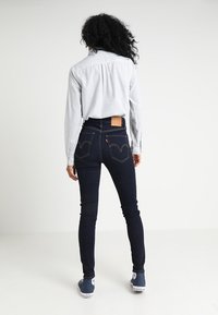 Jeans ajustados azul oscuro con cintura alta, costuras en contraste, bolsillos traseros con un parche de logo, combinados con una camisa azul claro.