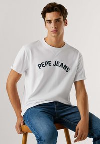 T-shirt blanc en coton avec un logo texte incurvé "PEPE JEANS" en bleu marine sur la poitrine, associé à un jean en denim bleu.