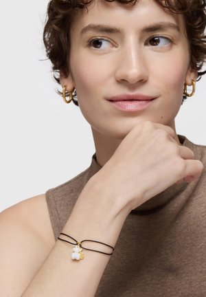 Pulsera de cordón negro con un conjunto de charms blancos y dorados. Modelo con top marrón sin mangas y pendientes de aro dorados.
