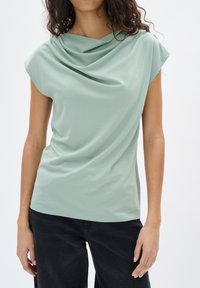 Top drappeggiato verde chiaro con maniche corte, tessuto morbido e una leggera lucentezza. Abbinato a pantaloni scuri dal taglio rilassato.