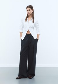 Chemise blanche à manches longues avec un design en V sur le devant, associée à un pantalon large noir et des chaussures marron foncé. Textures de tissu lisses.