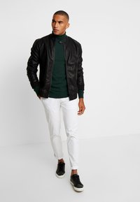 Jachetă bomber din piele neagră, pulover verde cu guler înalt, pantaloni albi și adidași negri. Jacheta are manșete și guler ribbenate, cu un design ajustat.