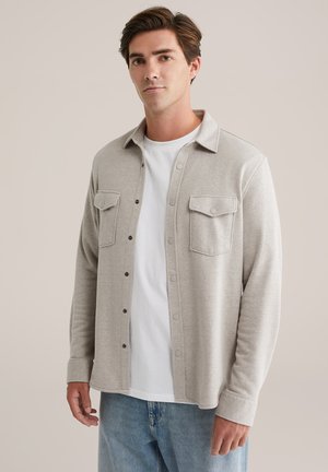 Chaqueta de camisa gris claro hecha de tela suave, con dos bolsillos en el pecho, cierre de botón y cuello. Usada sobre una camiseta blanca.