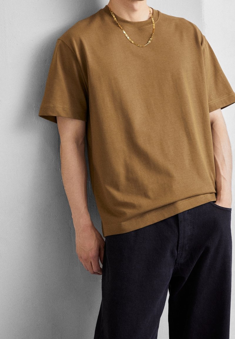 Homme portant un t-shirt marron à manches courtes, un pantalon noir et un collier en chaîne dorée, debout devant un mur texturé gris.