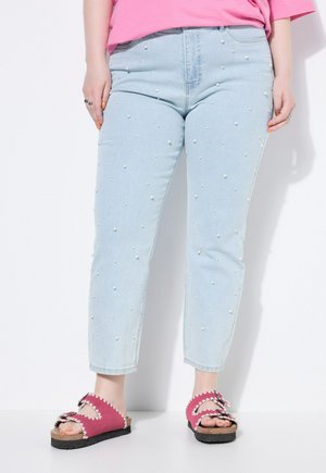 Jasnoniebieskie jeansy do kostek ozdobione małymi białymi perłami, zestawione z różowymi sandałami z falistymi krawędziami i klamrami na białym tle.