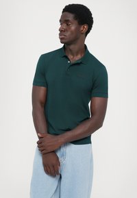 Polo de culoare verde închis, cu mâneci scurte, fabricat dintr-un material neted. Dispune de un guler cu nasturi și un broderie subtilă a logo-ului EA7 pe piept.
