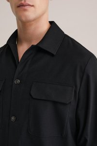 Camicia nera con bottoni e colletto, dotata di due tasche frontali. Il tessuto appare liscio e strutturato, con un design aderente.