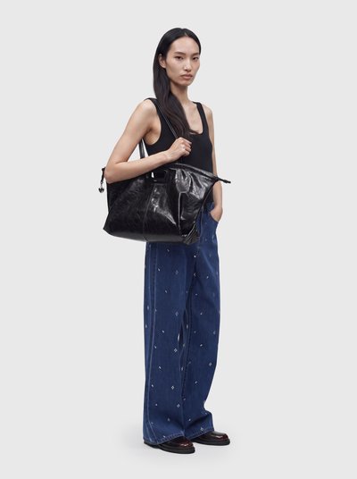 Femme aux longs cheveux foncés portant un haut noir sans manches et un pantalon large bleu, elle porte un grand sac à main en cuir noir sur l'épaule.