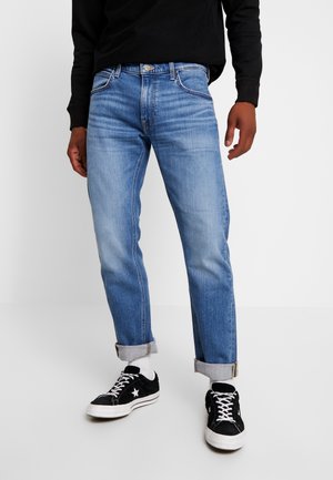 Mand iført lyseblå opkørte jeans, sort sweatshirt, hvide sokker og sorte lave sneakers med hvide snørebånd og stjerne-logo.