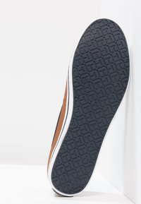 Brown leather sneaker with a patterned black rubber sole, white midsole, and navy accents along the side. Designed for casual wear.