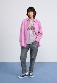 Camisa chaqueta de pana rosa sobre una camiseta gráfica gris con diseño de montañas. Combinada con jeans grises y zapatillas azul oscuro. Atuendo sencillo y casual.