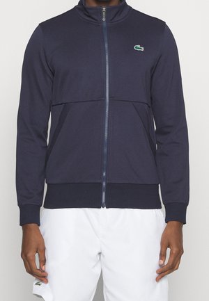 Giacca zip-up blu navy con colletto alto, polsini a coste e tasche. Realizzata in un tessuto testurizzato e presenta un piccolo logo verde sul petto.