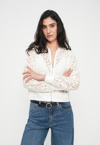 Veste bomber en dentelle blanche avec une fermeture éclair à l'avant, poignets et taille côtelés, présentant des motifs floraux complexes. Associée à un jean en denim bleu.