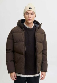 Giacca marrone imbottita con cappuccio e zip frontale, caratterizzata da un tessuto testurizzato. Indossata sopra un maglione scuro e stratificata con una camicia a righe.