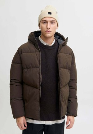 Giacca marrone imbottita con cappuccio e zip frontale, caratterizzata da un tessuto testurizzato. Indossata sopra un maglione scuro e stratificata con una camicia a righe.