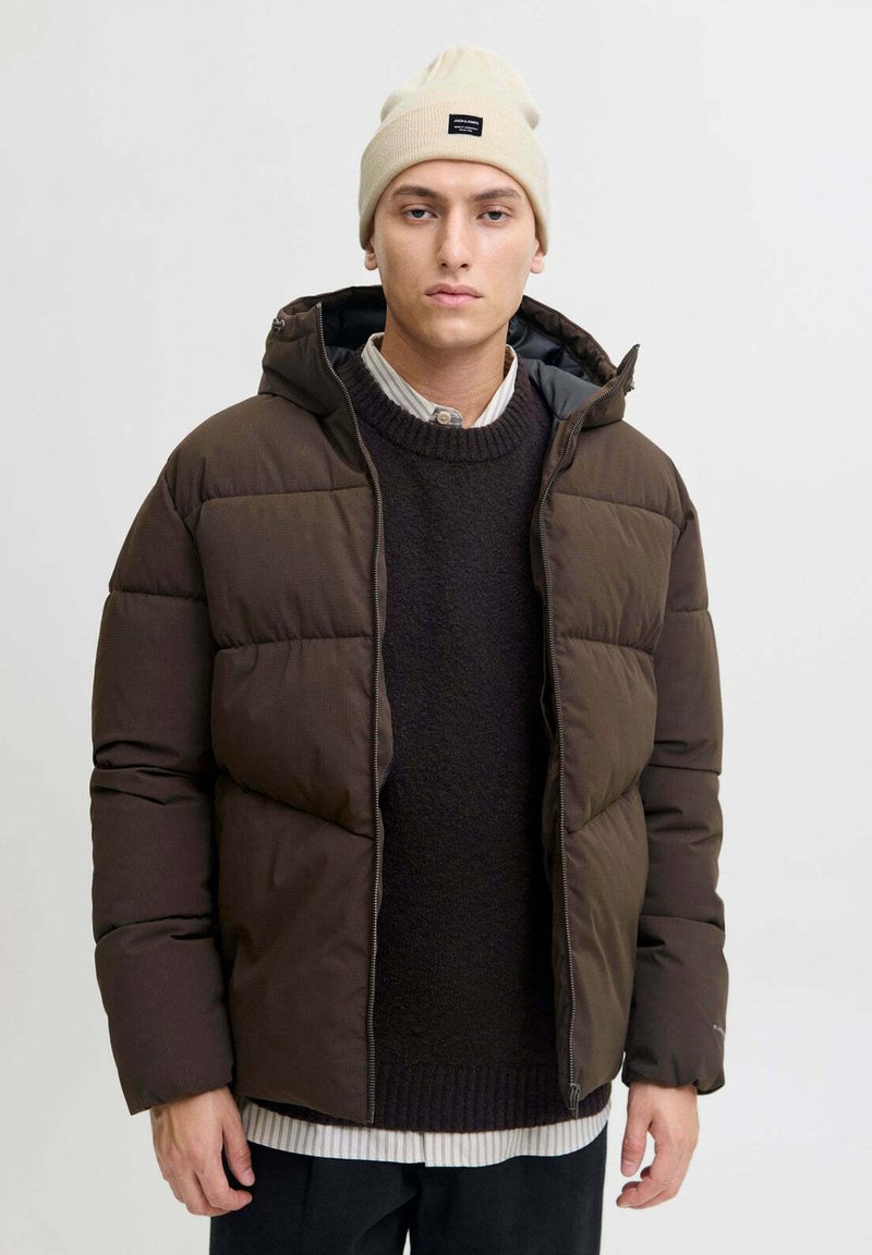 Giacca marrone imbottita con cappuccio e zip frontale, caratterizzata da un tessuto testurizzato. Indossata sopra un maglione scuro e stratificata con una camicia a righe.