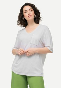 Camiseta blanca de cuello en V en tejido ligero con mangas cortas, que presenta un sutil detalle de bolsillo. Combinada con pantalones verdes texturizados.