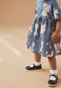 Modré šaty s květinovým a vílí potiskem, kratší rukávy a nařasený pas. Černé boty s bílými krajkovými ponožkami doplňují celý outfit.