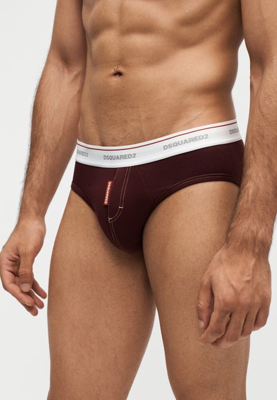 Torso masculin portant un slip bordeaux foncé avec une ceinture blanche portant l'inscription DSQUARED2, et une petite étiquette de marque sur le devant à gauche.