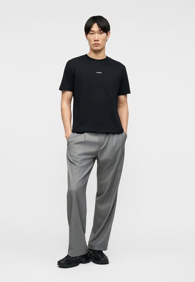 Νεαρός άνδρας στέκεται με τα χέρια στις τσέπες, φοράει μαύρο μπλουζάκι με μικρό κείμενο και γκρι παντελόνι ραμμένο με μαύρα παπούτσια.