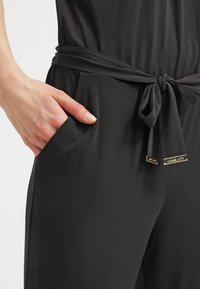 Main avec des ongles naturels placée dans une poche d’un pantalon noir avec une ceinture en tissu nouée et des étiquettes métalliques avec logo aux extrémités de la ceinture.