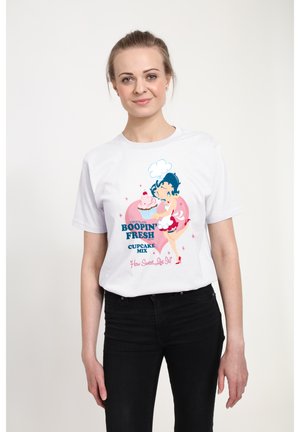 Hvid bomulds t-shirt med et farverigt cartoon-graphic af en karakter med blåt hår, der holder en cupcake, samt tekstelementer i pink og blå.
