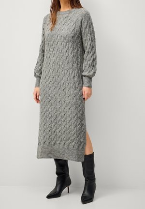 Kobieta w długiej, szarej, dzianinowej sukience z włóczki o splocie warkoczowym, z rękawami typu balon i czarnych botkach na obcasie z ostrym noskiem.