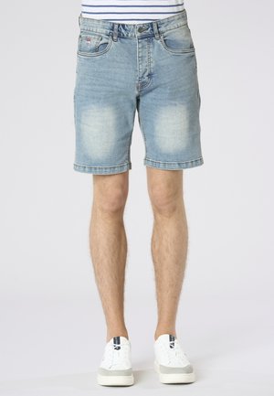 Shorts in denim azzurro chiaro indossati da una persona in piedi, abbinati a scarpe da ginnastica bianche e una camicia a righe, su uno sfondo semplice.