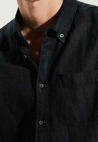 Gros plan sur une personne portant une chemise noire à col, déboutonnée, avec des boutons visibles et une poche poitrine avant.