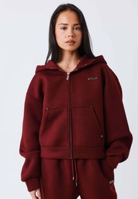 Sudadera con capucha con cremallera en burdeos, hecha de un tejido suave, con capucha, dos bolsillos frontales y logo en el pecho. Presenta un ajuste holgado y puños acanalados.