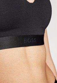 Juodas braletas, pagamintas iš lygaus audinio, su apvaliu kaklu ir plačia elastinga juosta, ant kurios tamsiomis raidėmis parašyta "BOSS".