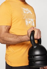 Oranžové bavlněné tričko s bílou grafikou na přední straně, která zobrazuje palmové listy a text "PUMA". V ruce je drženo černé kettlebell.