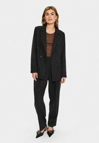 Donna che indossa un completo grigio scuro con blazer e pantaloni a vita alta, top marrone e scarpe nere con punta e cinturini alla caviglia, in piedi davanti a uno sfondo bianco.