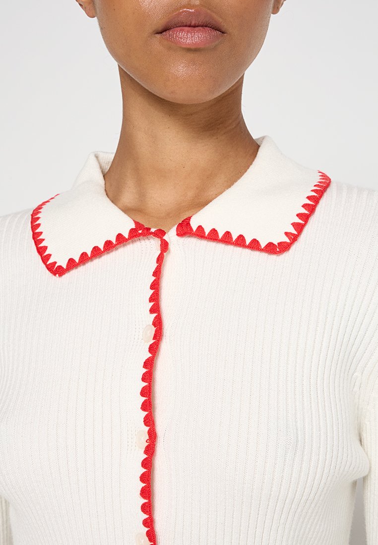 Weißer gerippter Pullover mit einem gewellten roten Kragen und Knopfverschluss, der ein strukturiertes Design und kontrastierende Farbdetails hervorhebt.
