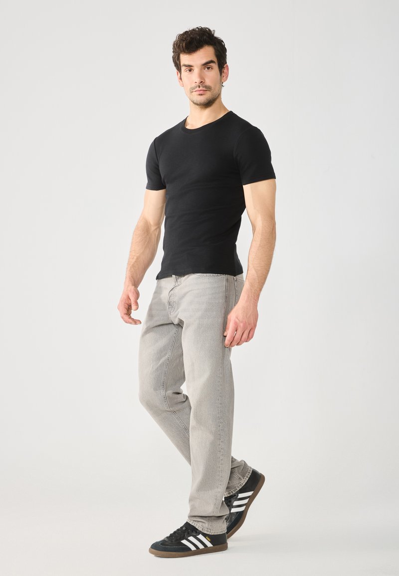 Schwarzes T-Shirt mit kurzen Ärmeln, hellgraue Jeans und schwarze Sneakers mit weißen Streifen. Sleek Fit und glatte Textur. Stehende Pose vor neutralem Hintergrund.