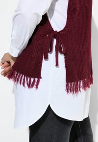 Chaleco de punto burdeos con dobladillo de flecos, que cuenta con un cierre trasero atado. Se lleva sobre una camisa blanca con dobladillo curvado y jeans negros.